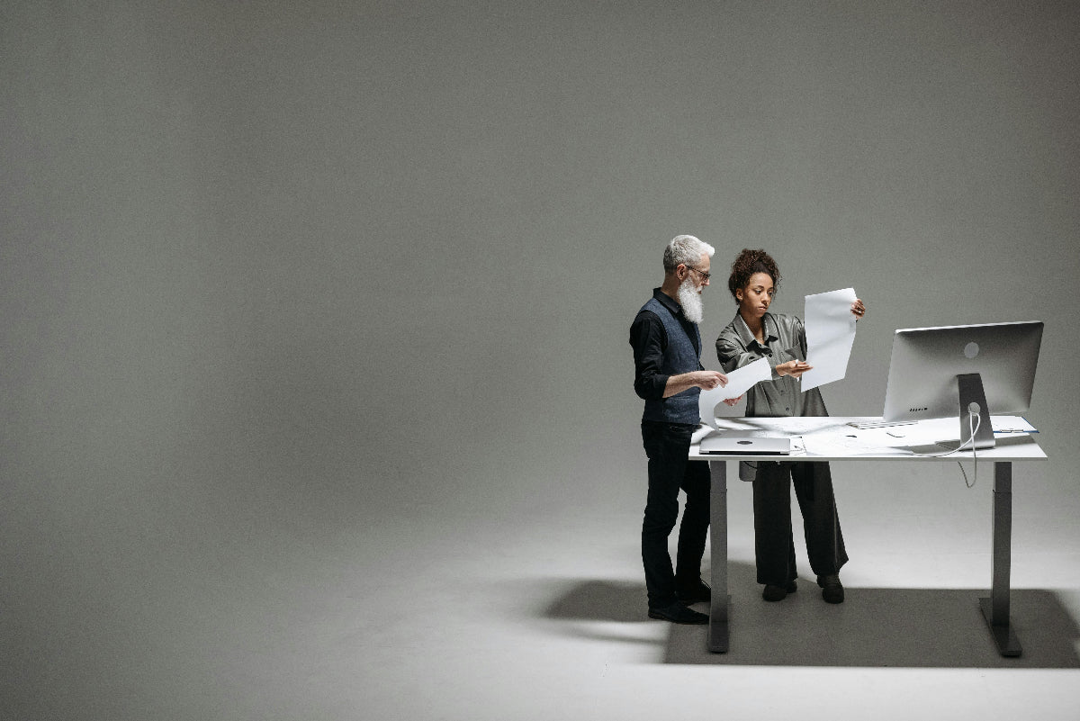 Two people working at a desk with a computer and documents on a gray background