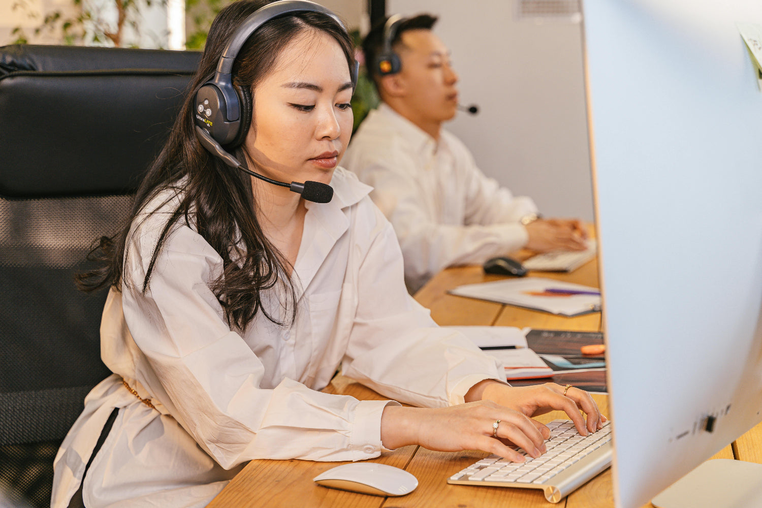 Two people working at a desk with computers and headsets.